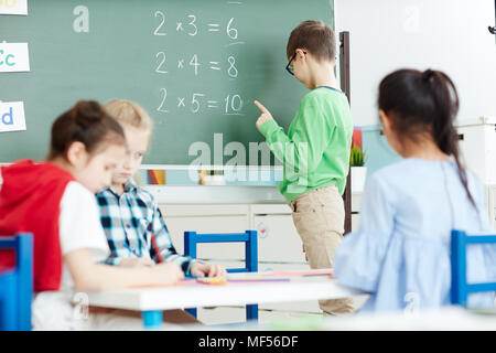 Smart little schoolboy standing by blackboard in classroom and doing multiplication sums on math class Stock Photo