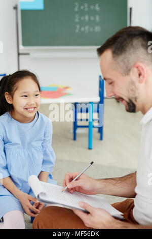Male primary school teacher writing something on clipboard while giving private lesson to cheerful Asian girl sitting next to him Stock Photo
