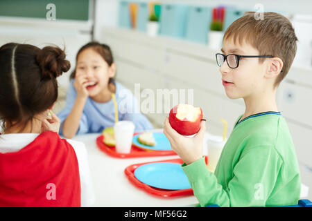 Caucasian boy in glasses eating apple while having lunch with classmates in elementary school cafeteria Stock Photo