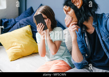 Father wearing monkey mask looking at son using smartphone at home Stock Photo