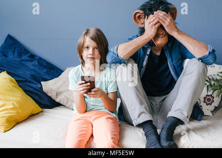 Father wearing monkey mask sitting next to son using smartphone at home Stock Photo