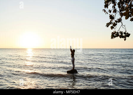 Young woman practicing yoga on a rock in the sea at sunset Stock Photo