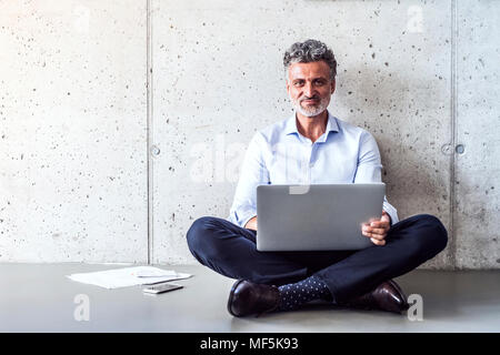 Portrait of confident mature businessman sitting on the floor using laptop Stock Photo