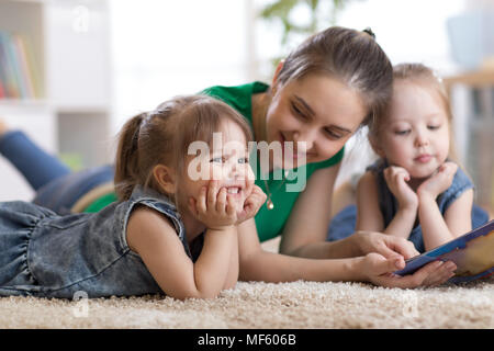 Children laughing and having fun reading stories with their mother laying on the floor at home Stock Photo