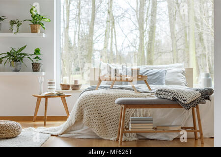 Blanket on wooden bench with books in front of bed in cozy bedroom interior with plants Stock Photo