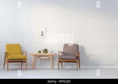 Wooden table between yellow and grey armchair against white wall with poster in living room interior Stock Photo
