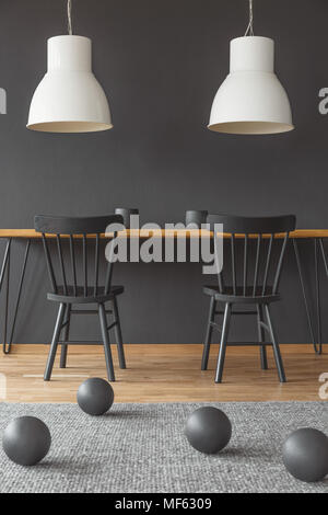 Two, white lamps hanging above a wooden dining table in black room interior with balls decorations on the floor Stock Photo