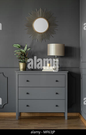 Gold round mirror on grey wall above cabinet with lamp and plant in living room interior Stock Photo
