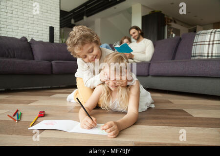 Siblings playing on floor while parents reading book on sofa Stock Photo