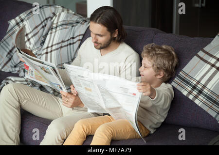 Cute kid son reading newspaper sitting on sofa with dad Stock Photo