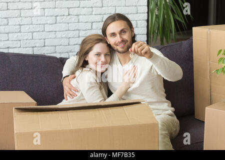 Happy couple holding keys looking at camera sitting on couch  Stock Photo