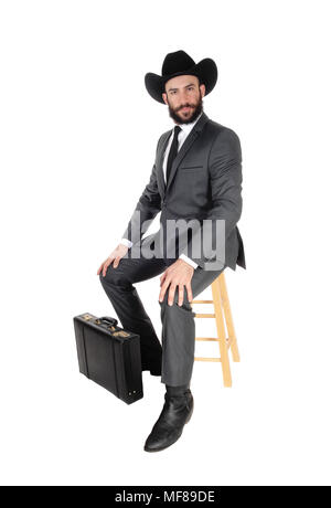 A young handsome business man wearing a suit and cowboy hat sitting on a bar chair with his briefcase, isolated for white background Stock Photo