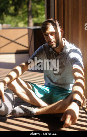 Young man in sun rays sitting on terrace and meditating, wearing shorts. Stock Photo