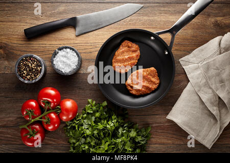Raw meatballs in a black frying pan on a wooden table. Tomatoes, meat knife, napkin, parsley, black pepper and meat salt props are on the table. Stock Photo