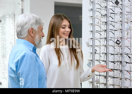 Sideview of smiling assistant showing eyeglasses to old patient. Advising which optical glasses are better. Tranparent stand with glasses set in front of white wall on background. friendly and kind.