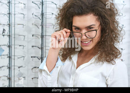 Charismatic, confident oculist wearing white uniform. Woman wearing fashionable lenses. Doctor is standing in front of transparent stand with stylish, modern optical eyeglasses. Stock Photo