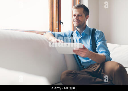 Handsome man sitting on sofa at home, using a digital tablet Stock Photo