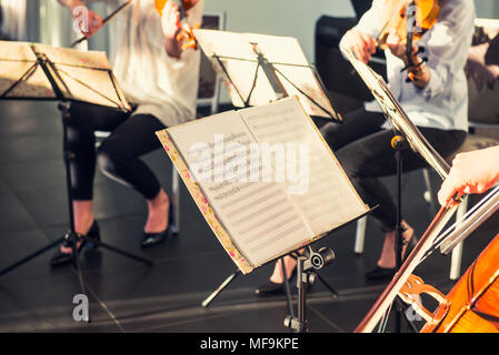 Selective Focus on Music note sheets on stand with background of playing cellists and violinists band on event in bright day sunlight. Commercial musi Stock Photo