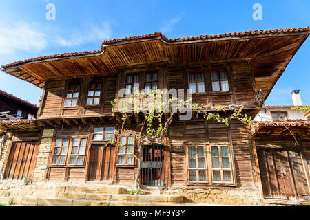 Low angle view of the front side of old traditional rustic house made of wood and covered by grape vine with eaves on top against blue sky, Zheravna,  Stock Photo