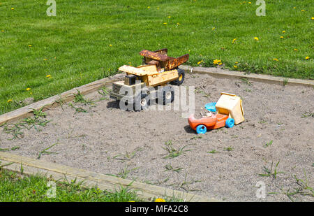 Old worn toys in a sandbox surrounded by green grass wtih some dandelions. Stock Photo