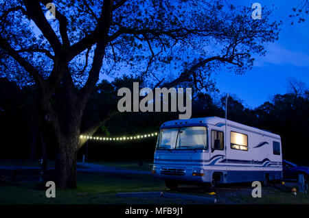 A large motorhome sits under a huge pecan tree, lit up as night falls on the campsite. Stock Photo