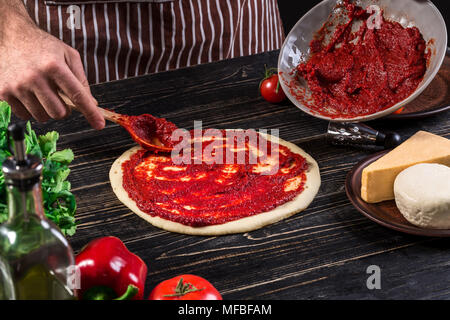 A male hand spreading tomato puree on a pizza base with spoon on an old wooden background Stock Photo