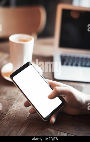 Young man with tattoo holding mobile phone and working on laptop in co-working. Mans hand and phone are on foreground and on focus. Stock Photo