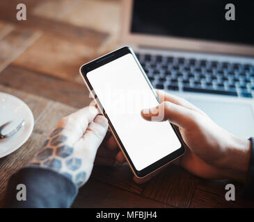 Young man with tattoo holding mobile phone and working on laptop in co-working. Mans hand and phone are on foreground and on focus. Stock Photo