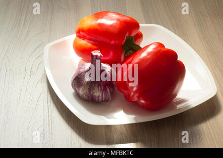 Two red bell peppers and a head of blue garlic lie on a white plate Stock Photo