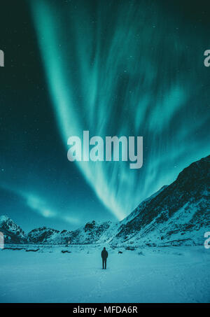 Young man is standing on a remote beach watching the Northern Lights dance above mountains in winter, Lofoten Islands archipelago, Norway, Scandinavia Stock Photo