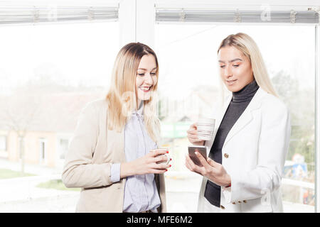 Two female collegues standing next to each other, looking at mobile phone and discussing about work while drinking coffee in an office. Stock Photo