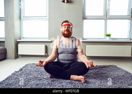 A funny fat bearded man in the headphones does yoga. Stock Photo