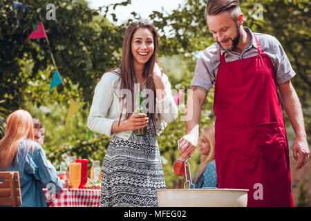 Cheerful young couple standing by a grill, the woman holding a green bottle of beer, the man holding cooking tongs at a summer garden party