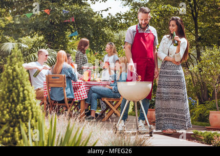 A couple standing by a grill, the man turning around a sausage, the woman with a beer bottle in her hand, with other people by a table enjoying their 