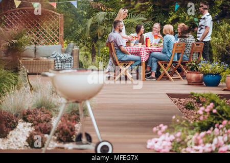 Wooden patio in the garden with a grill standing in the front and happy young people gathered around a table full of food during summer brake meeting Stock Photo