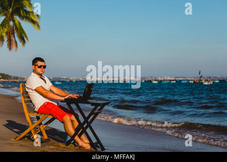 Man working with a laptop, on a hammock in the beach. Concept of digital nomad, remote worker, independent location entrepreneur. Stock Photo