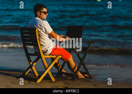Man working with a laptop, on a hammock in the beach. Concept of digital nomad, remote worker, independent location entrepreneur. Stock Photo