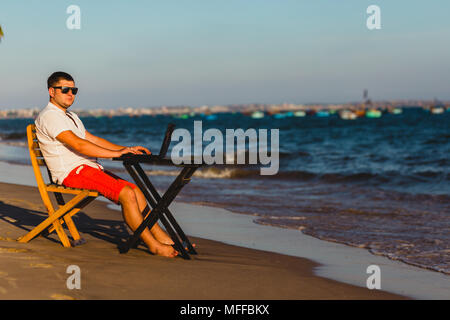 Man working with a laptop, on a hammock in the beach. Concept of digital nomad, remote worker, independent location entrepreneur. Stock Photo