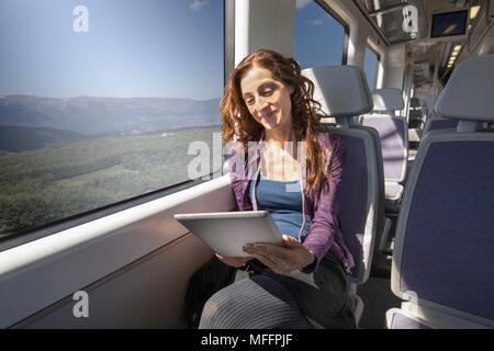 red hair smiling woman dressed in purple and blue, traveling by train sitting reading digital tablet or ebook Stock Photo
