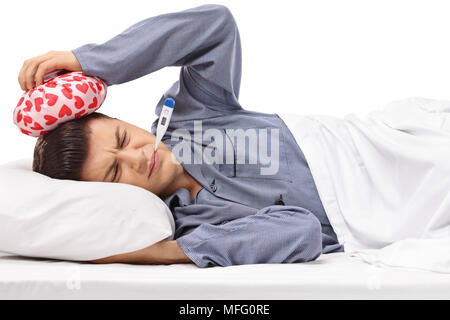 Sick teenager lying in bed holding an icepack on his head and a thermometer in his mouth isolated on white background Stock Photo