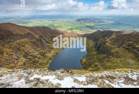 Aerial view from the top of the mountain ridge overlooking blue lake and glacial, horseshoe valley in Southern Ireland. Snow, farmlands and  coast. Stock Photo