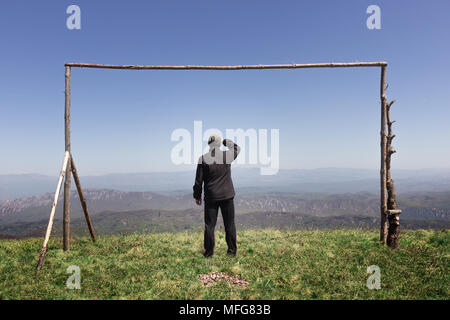 A man looking at a distance from a vantage point, standing in the middle of wooden frame, with a hat and his hand above his eyes to cover the sun Stock Photo