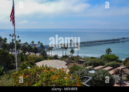 San Clemente Pier, Southern California, on a clear day, American US Flag on flagpole in the foreground, blue sky with clouds, Pacific Ocean shoreline Stock Photo