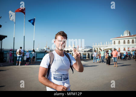 Young trendy man stands on the city square and shows gesture. Serious guy on background of ferry terminal and local people. Stock Photo