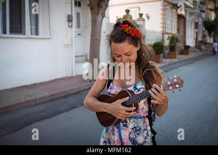 Smiling woman with roses in hairstyle holds in hands musical instrument. Pretty girl walks on the street and plays on ukulele. Stock Photo