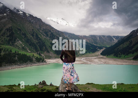 Handsome young woman is standing on the hill with view on the azure lake and green mountains with snowy tops and dramatic clouds. Stock Photo
