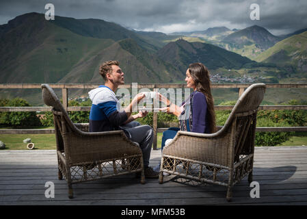 Young couple sits together on the terrace with cups of coffee in the morning. Man sends air kiss to girlfriend. Travelers having breakfast with beauti Stock Photo