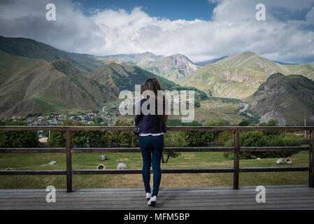Young woman stands back on observation deck with amazing view on the mountains and small village in valley. Stock Photo