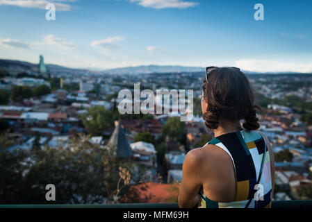 Young unrecognizable woman is standing back at observation deck. View from the hill on old town of Tbilisi. Cityscape in the evening. Stock Photo