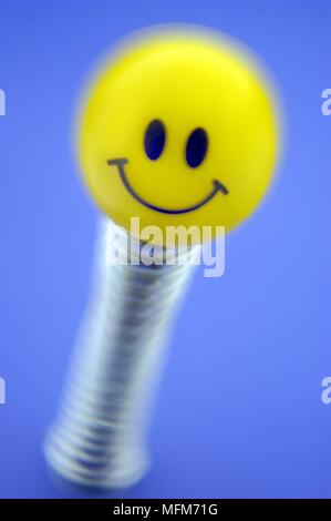 A happy smiling face painted on a yellow rubber ball balanced on a shiney chromed spring ready to bounce .   Shot on an electric blue background.    B Stock Photo
